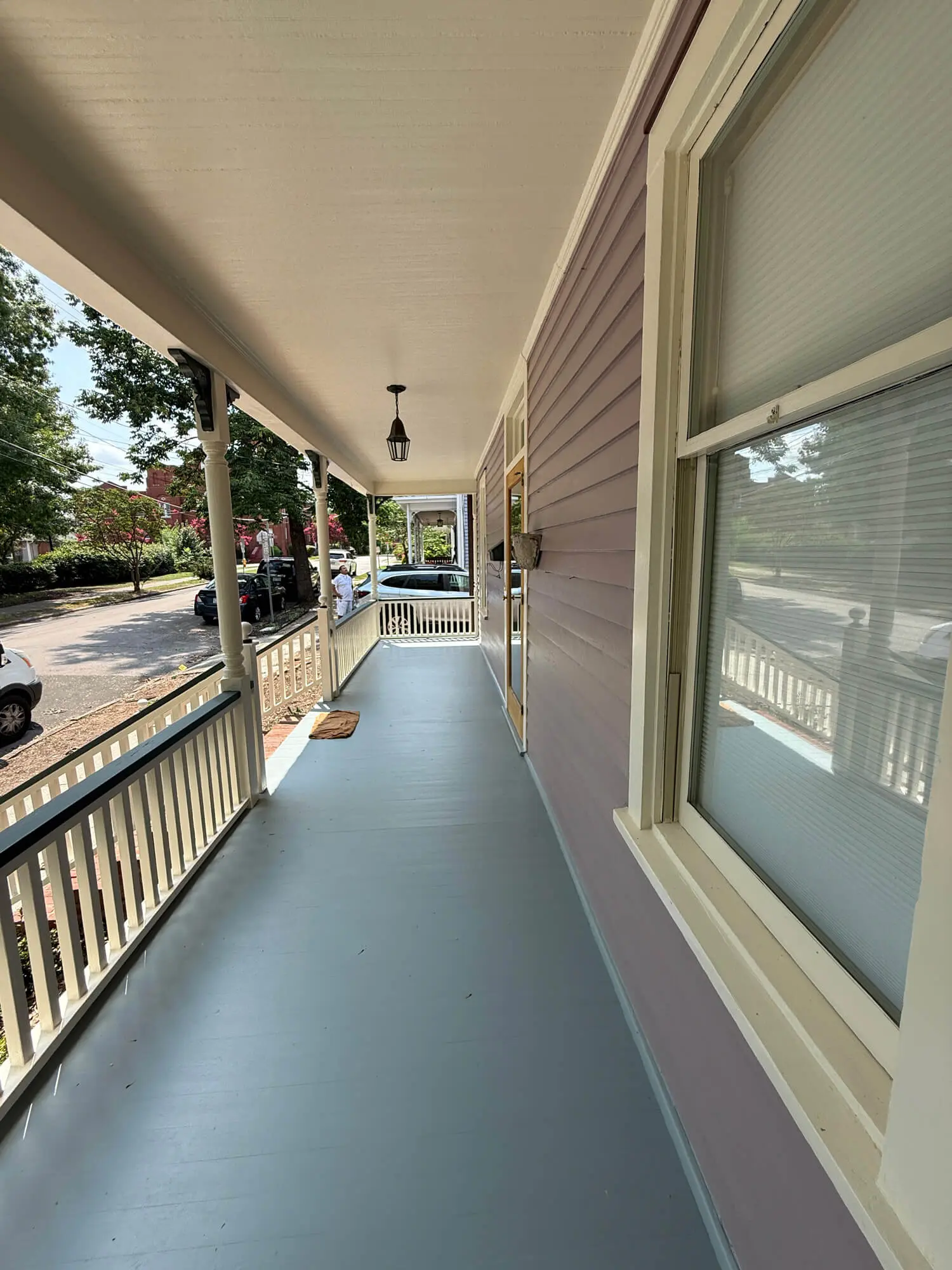 A freshly painted gray porch with white railings and columns, attached to a purple house with a window and door, overlooking a street with parked cars and trees.
