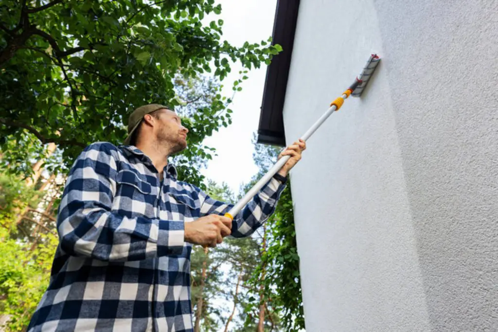 Painter rolling fresh paint onto the exterior wall of a house with green trees in the background