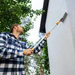 Painter rolling fresh paint onto the exterior wall of a house with green trees in the background